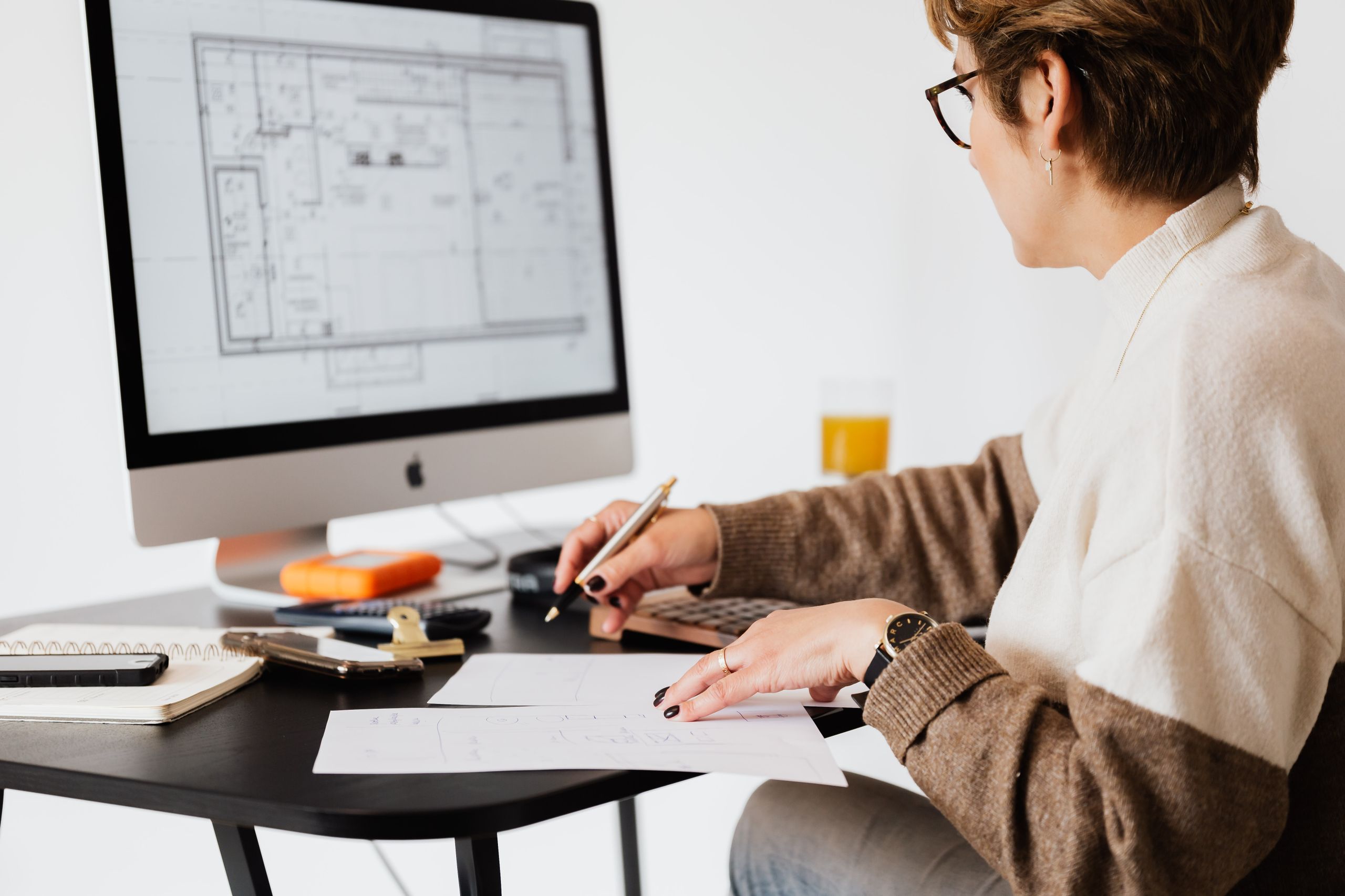 Side view of crop elegant female estate agent looking at floor plan on computer screen and noting down details on paper in light modern office